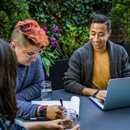 man in orange crew neck shirt using laptop beside two people