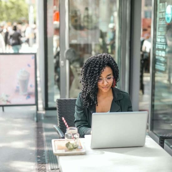 ethnic young woman using laptop while having tasty beverage in modern street cafe