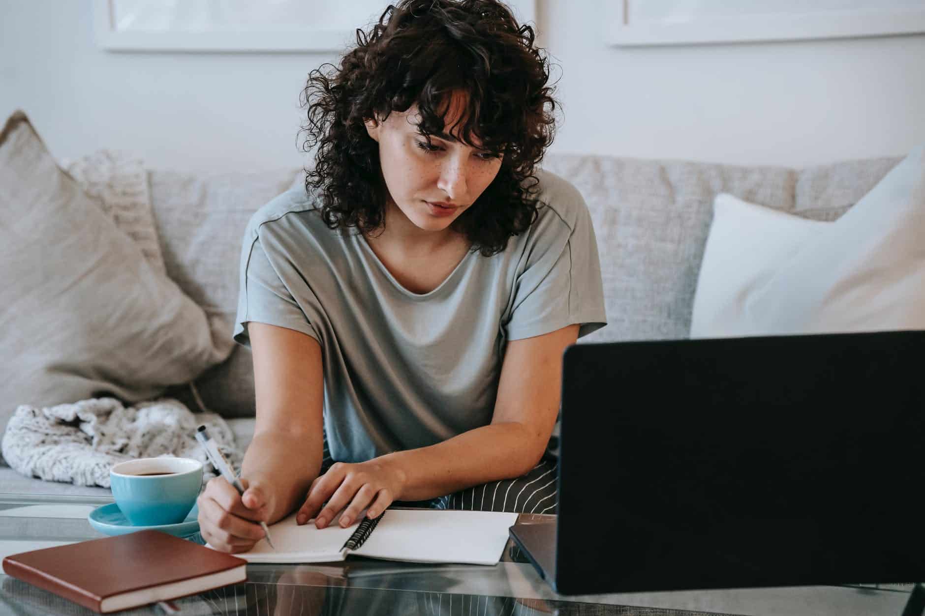 serious young lady writing in notebook during online studies on laptop at home