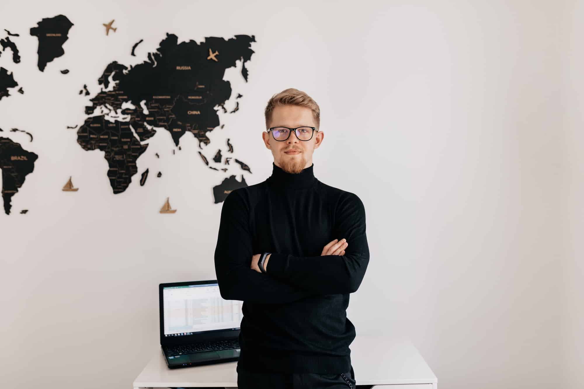indoor-portrait-handsome-blond-man-wearing-spectaculars-black-pullover-posing-white-wall-with-world-map-laptop-desktop