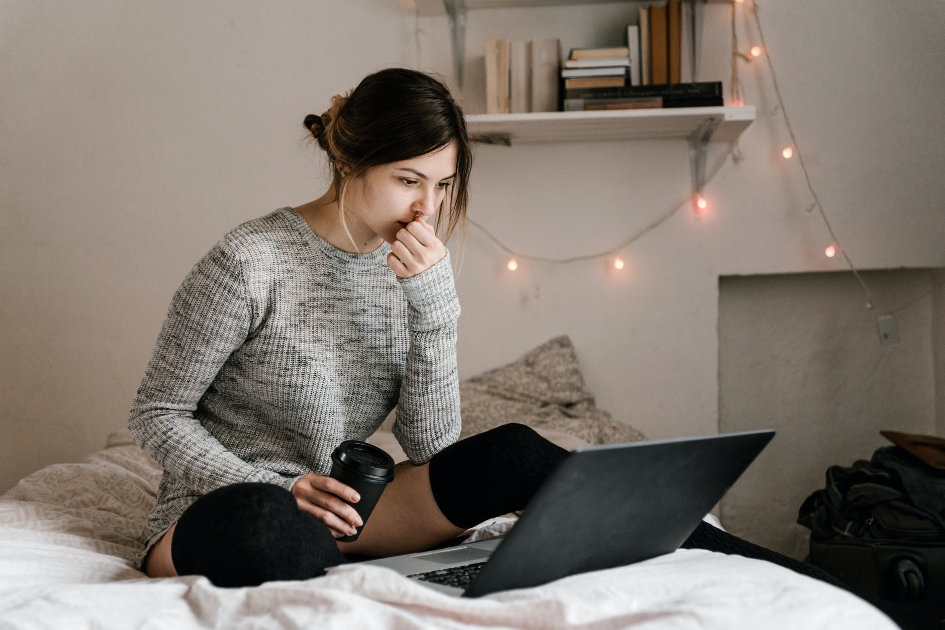 confused woman in gray sweater sitting on bed using macbook